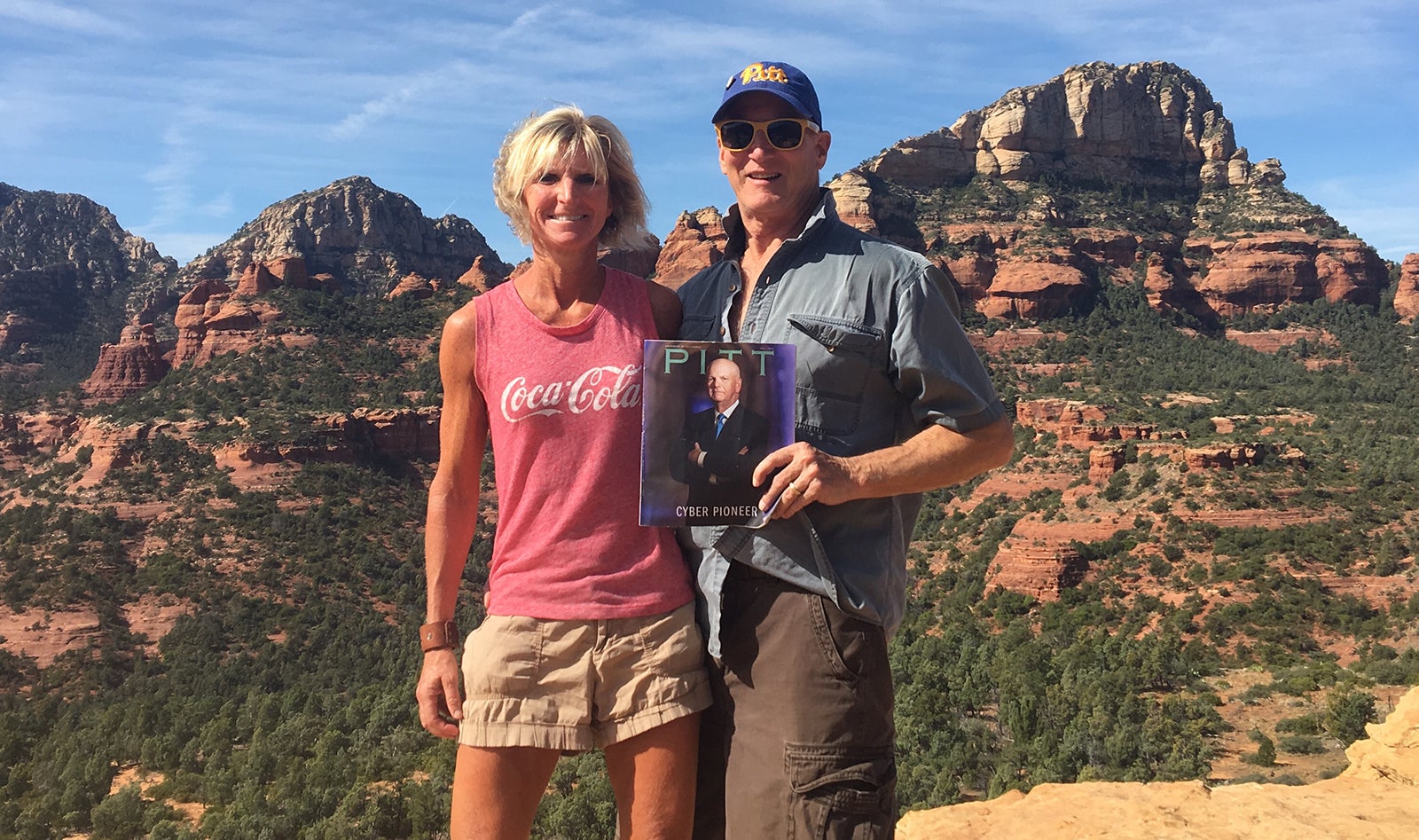 blond-haired woman in shorts and Coca-Cola tank top and man in short-sleeves and Pitt script hat hold Fall 2019 issue of Pitt Magazine in front of rocky landscape