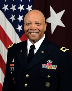 U.S. flag in background, wearing in formal uniform with tie, U.S. pin on one lapel and an American flag pin on the other lapel