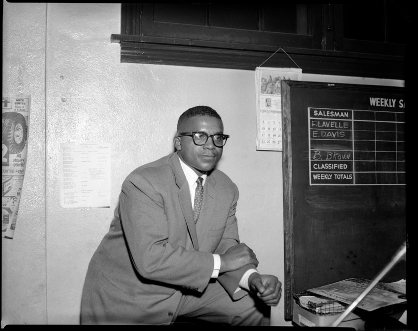 Young black man in suit and tie leans on knee, leg propped up, wears dark rimmed glasses in an interior space with chalkboard, calendar and tire advertisement