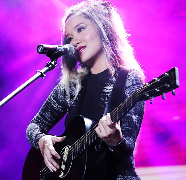 woman in front of microphone, playing black acoustic guitar, magenta background