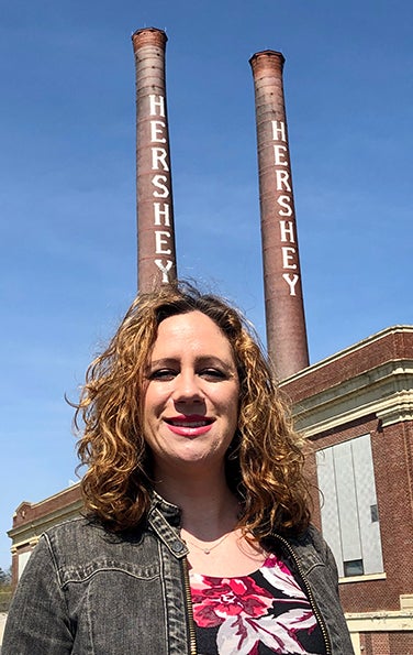 Tara Kennedy Griffiths in floral shirt and jean jacket in front of old Hershey factory stacks