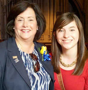 Karen, wearing a blue blazer and patterned blouse with a yellow rose pinned to her lapel, with daughter Maggie, in a red dress, at