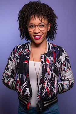 Leah wears her hair naturally curly, a multi-colored jacket, long gold necklace, white t-shirt and jeans, and stands against a purple backdrop.
