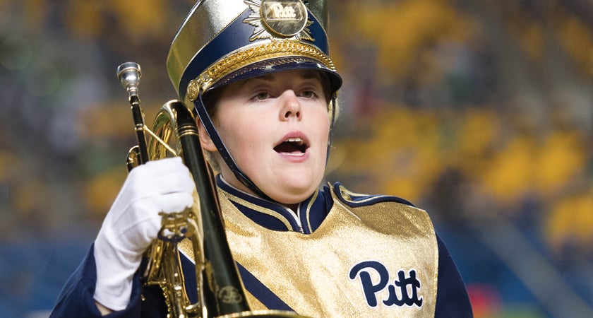 A woman opens her mouth to sing while wearing a gold and blue band uniform and holding a mellophone on her shoulder.